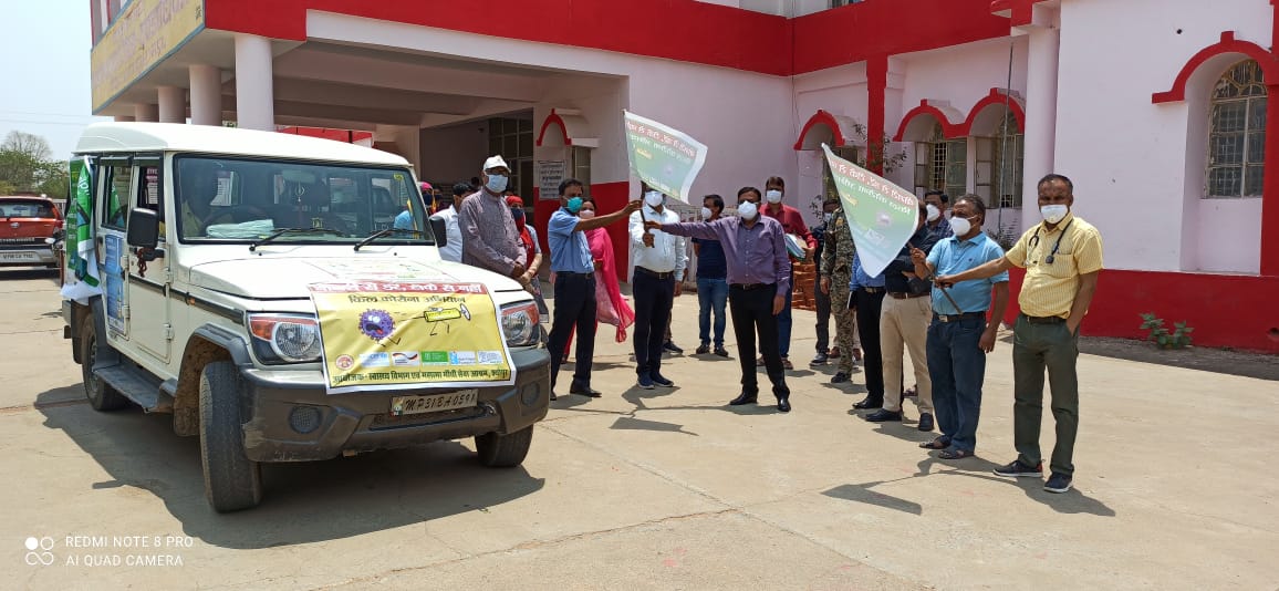 A group of people are standing near a vehicle wearing masks and holding banners. The vehicle is a jeep covered in banners with information about a COVID hotline and about COVID vaccines. The setting is outside the MGSA Joura Center in India.