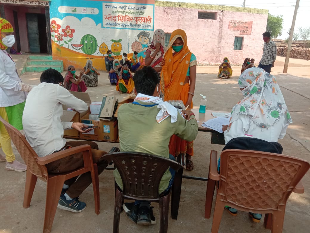 3 people sit at a table outsidie with boxes and papers, while a line up of masked people, mostly women wait for medicine. Photo is set in an outdoor location in India